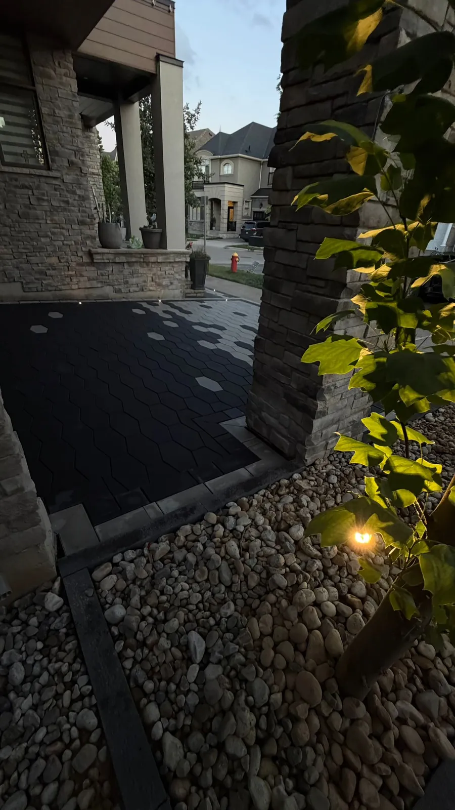 Dark hexagon paver driveway courtyard beside a stone-clad front porch at dusk