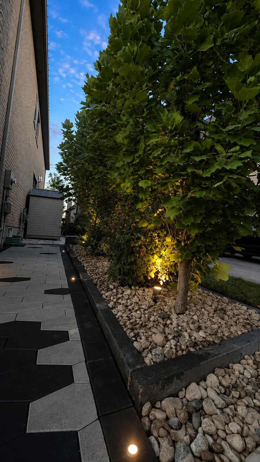 Side-yard hexagon paver walkway with a river-rock planting bed and uplighting at dusk